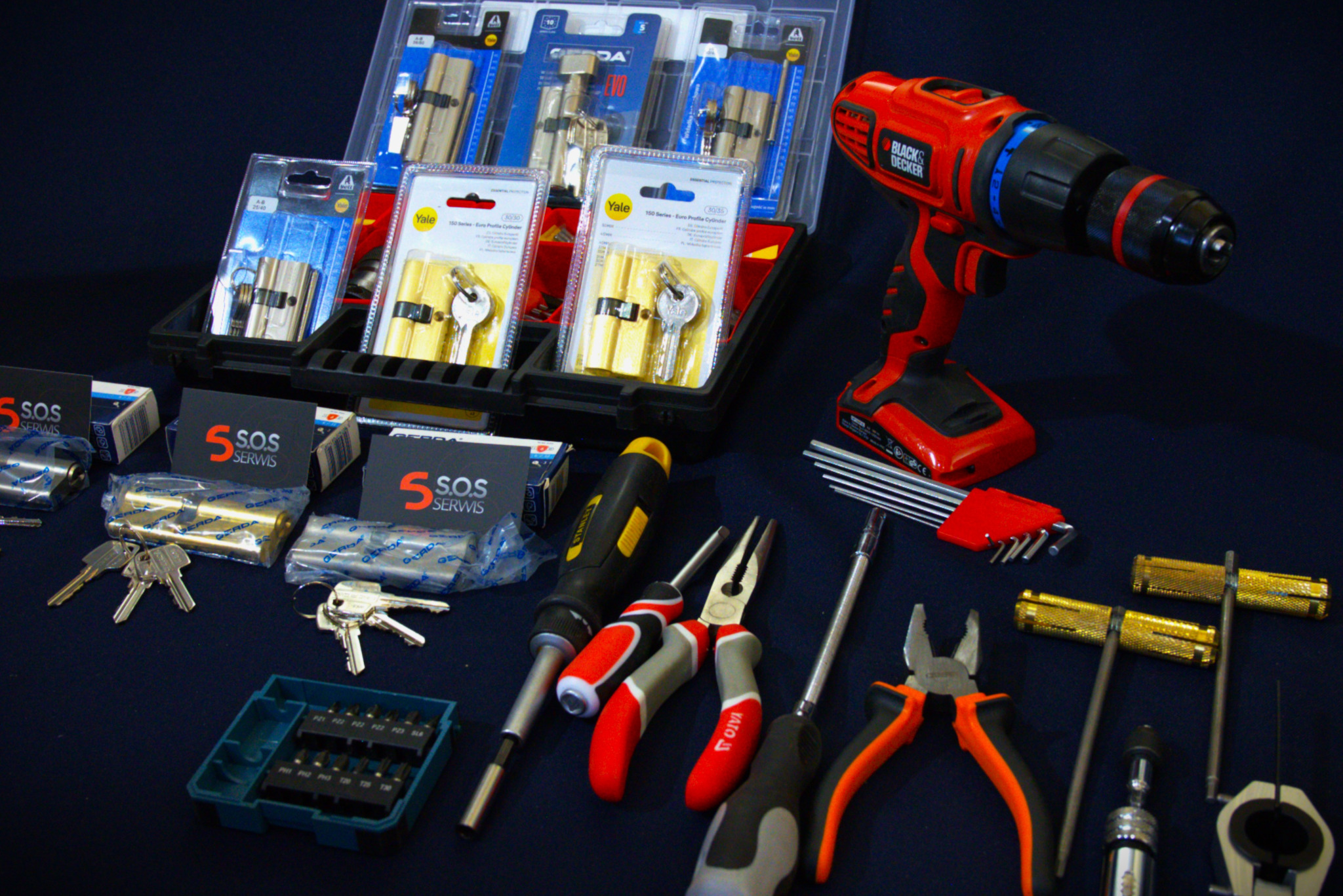 Zestaw narzędzi: wiertarka, śrubokręty, kombinerki, klucze, wkładki do zamków Gerda i Yale, oraz wizytówki S.O.S Serwis, na ciemnym tle.