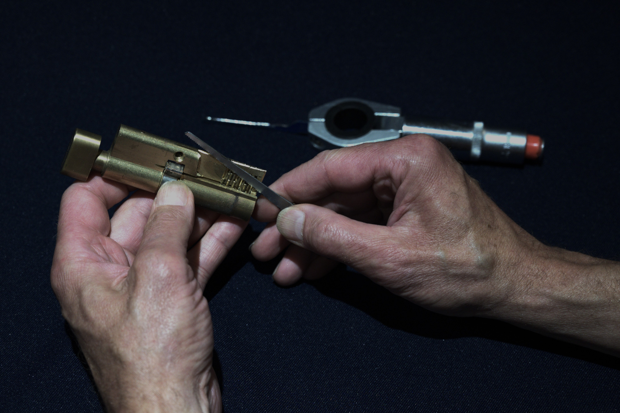 Otwieranie zamka wytrychem przez osobę na ciemnym tle. Widoczne narzędzia i zamek w kolorze złotym. Precyzyjna praca manualna.