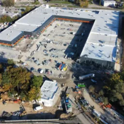 Widok z lotu ptaka na plac budowy hali w kształcie litery U z pomarańczowymi akcentami, z widocznymi fundamentami i maszynami budowlanymi w jesiennej scenerii.