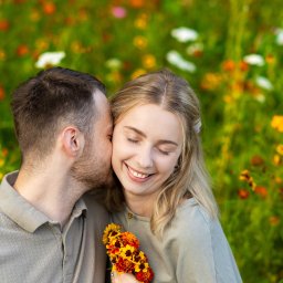 FOTKI NA ZAWSZE - Uśmiechnięta blondynka z bukietem polnych kwiatów, całowana w policzek przez mężczyznę na tle łąki pełnej różnobarwnych kwiatów.