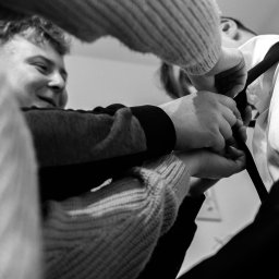 FOTKI NA ZAWSZE - Czarno-białe zbliżenie na mężczyznę w trakcie ubierania, prawdopodobnie muchy, z pomocą drugiej osoby. Widoczne dłonie i fragmenty ubioru.