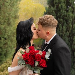 FOTKI NA ZAWSZE - Pocałunek pary młodej w plenerze. Panna młoda z czarnymi włosami trzyma bukiet czerwonych róż. Pan młody w czarnym garniturze. Tło stanowią zielone drzewa.