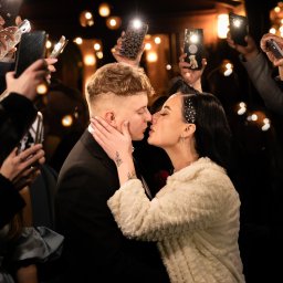 FOTKI NA ZAWSZE - Pocałunek pary młodej otoczonej przez gości robiących zdjęcia telefonami z włączonymi lampami błyskowymi, tworząc efektowne oświetlenie. Ciemne tło z rozproszonymi światłami.