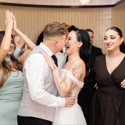 FOTKI NA ZAWSZE - Uśmiechnięta para młoda w objęciach, otoczona gośćmi weselnymi podczas radosnego świętowania. Panna młoda z tatuażami i ozdobami we włosach.