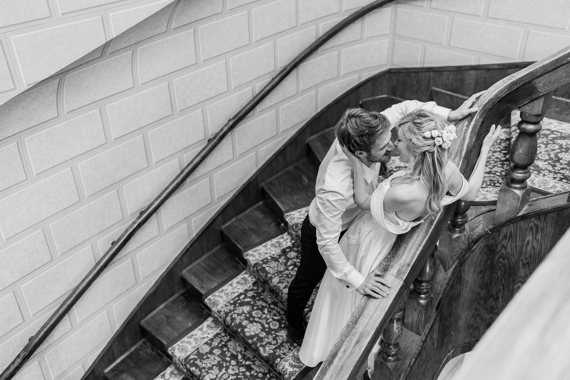Czarno-białe zdjęcie z góry: para młoda na drewnianych schodach z wzorzystym dywanem, pocałunek, kwiaty we włosach panny młodej, elegancka balustrada.