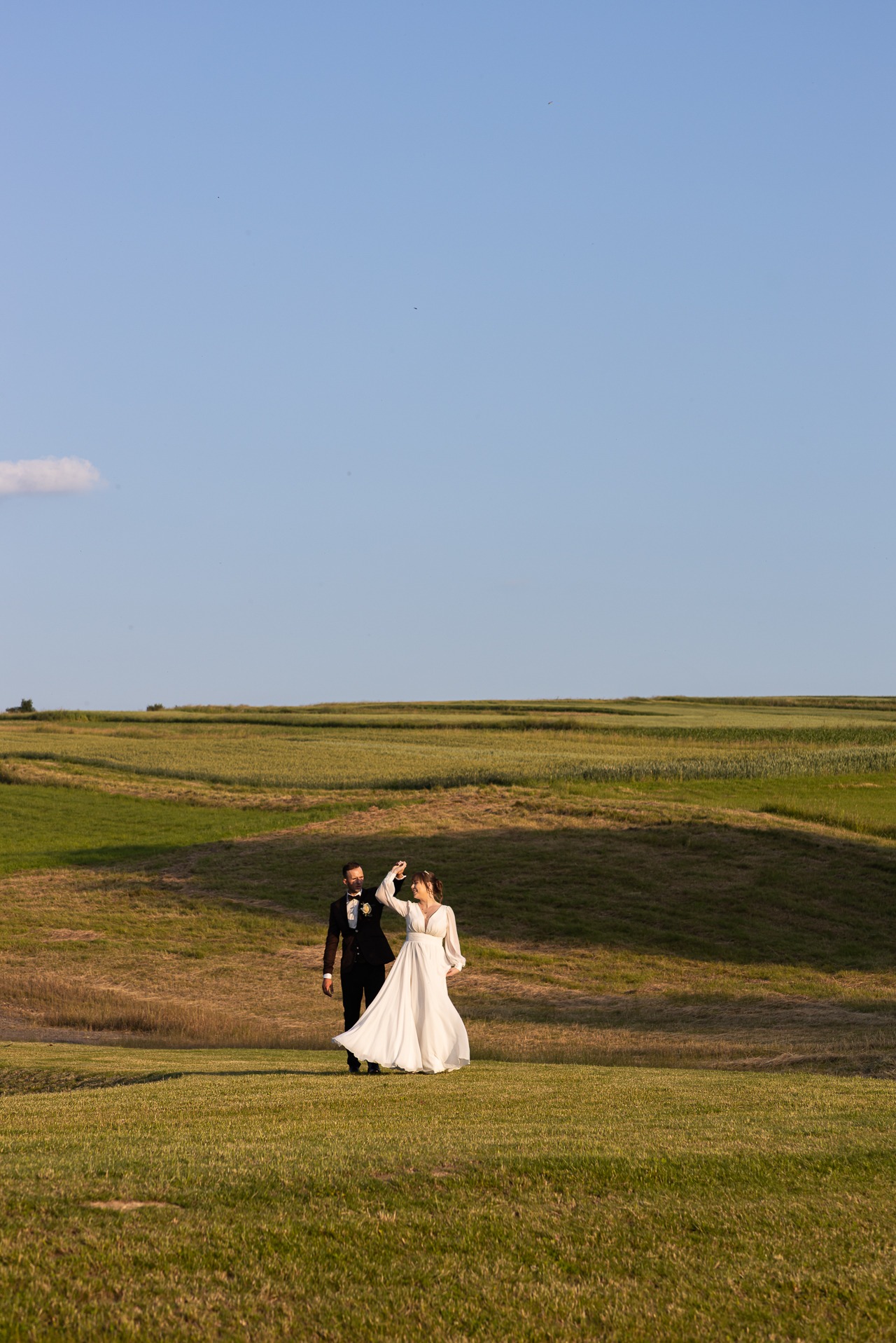 Para młoda na tle zielonych wzgórz i błękitnego nieba. Panna młoda w białej sukni z długimi rękawami, pan młody w czarnym smokingu z muszką. Romantyczny taniec.