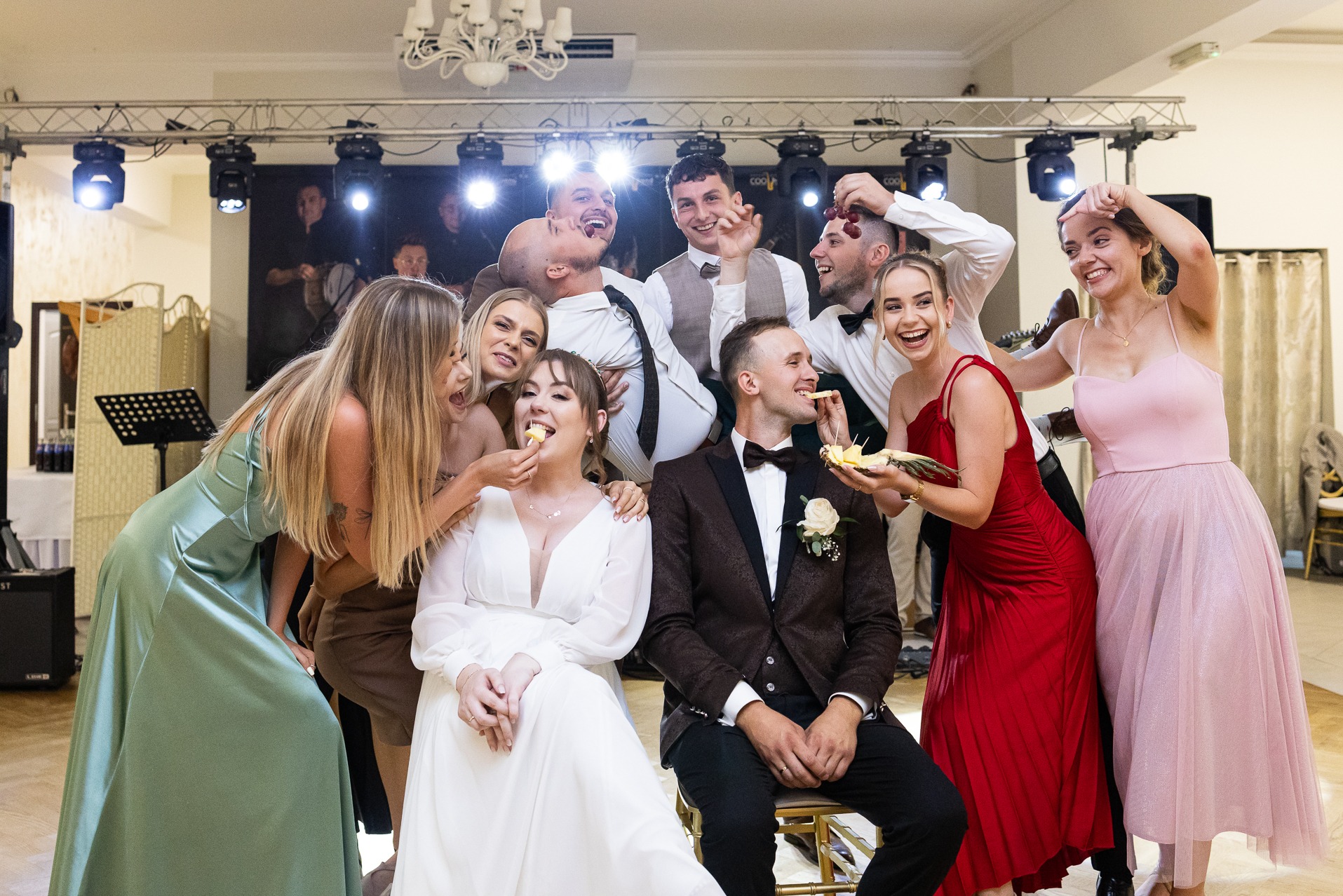 Radosna para młoda siedzi otoczona gośćmi weselnymi, którzy karmią ich kawałkami ananasa i winogron na tle sceny z oświetleniem i grającym zespołem.