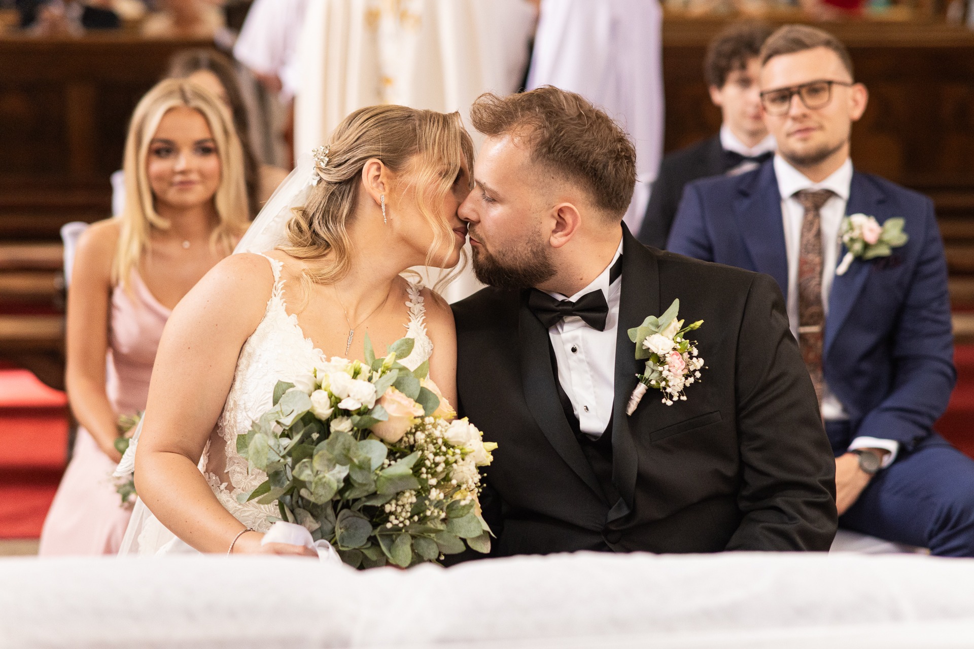 Pocałunek młodej pary w kościele. Panna młoda w koronkowej sukni trzyma bukiet. Pan młody w czarnym smokingu z muszką. W tle goście weselni.