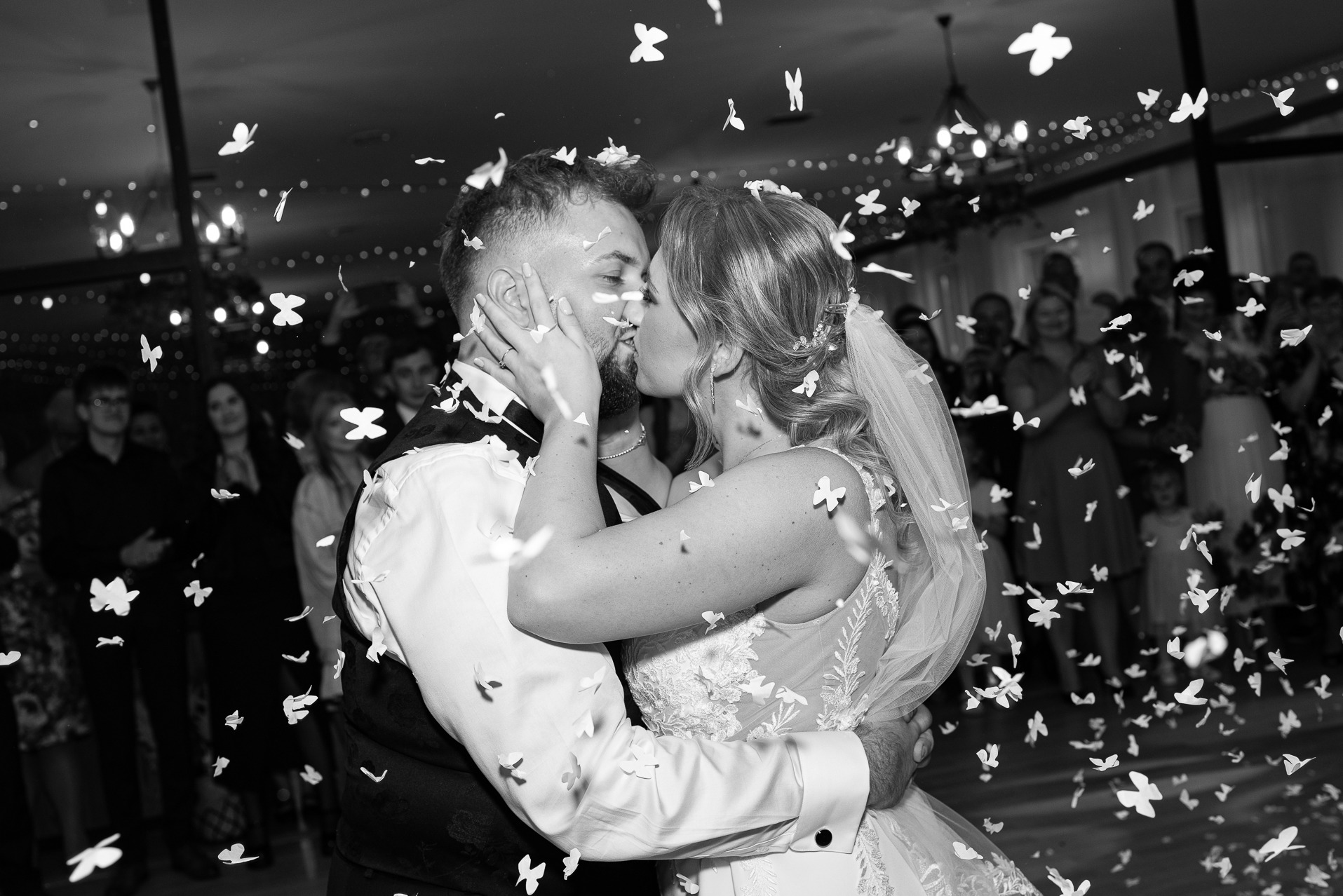 Czarno-białe ujęcie całującej się pary młodej w otoczeniu konfetti w kształcie motyli, z gośćmi w tle i dekoracyjnymi światłami.