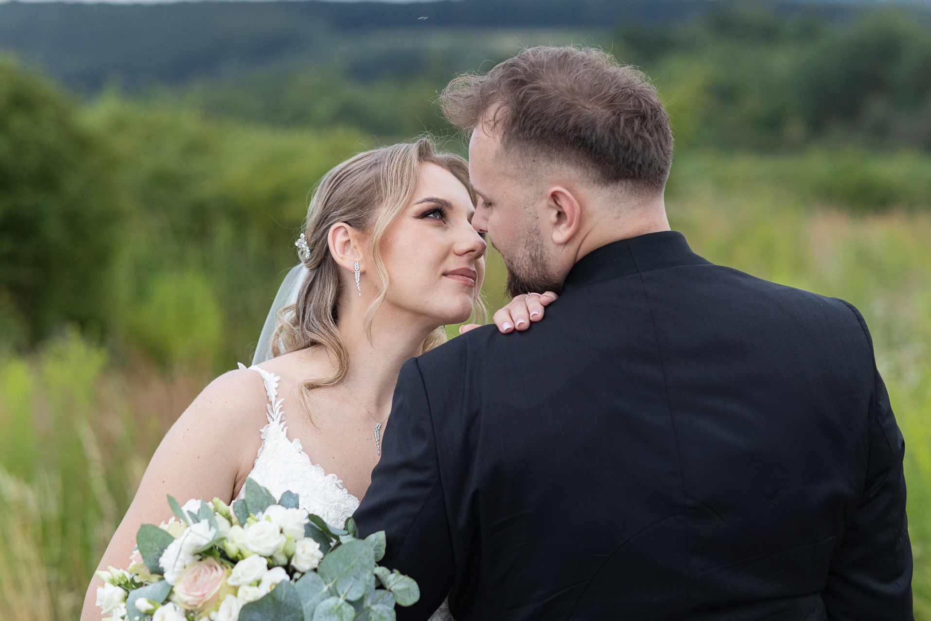 Para młoda w plenerze: panna młoda z bukietem spogląda na pana młodego w czarnym garniturze, zieleń w tle. Romantyczny moment przed obiektywem.