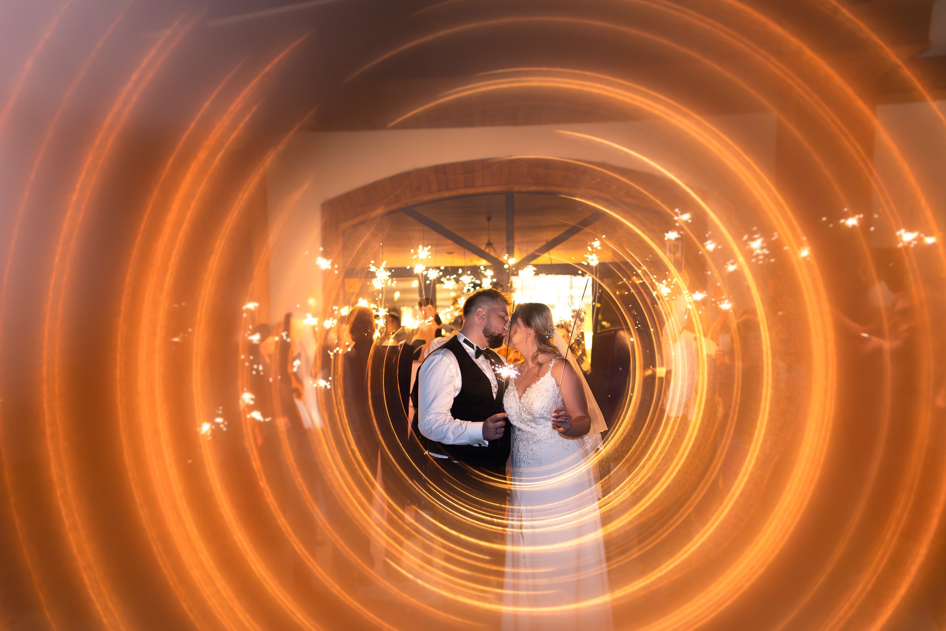 Pocałunek pary młodej otoczonej iskrami zimnych ogni na tle ceglanej ściany z oknem w kształcie łuku. Ujęcie z efektem świetlnym.