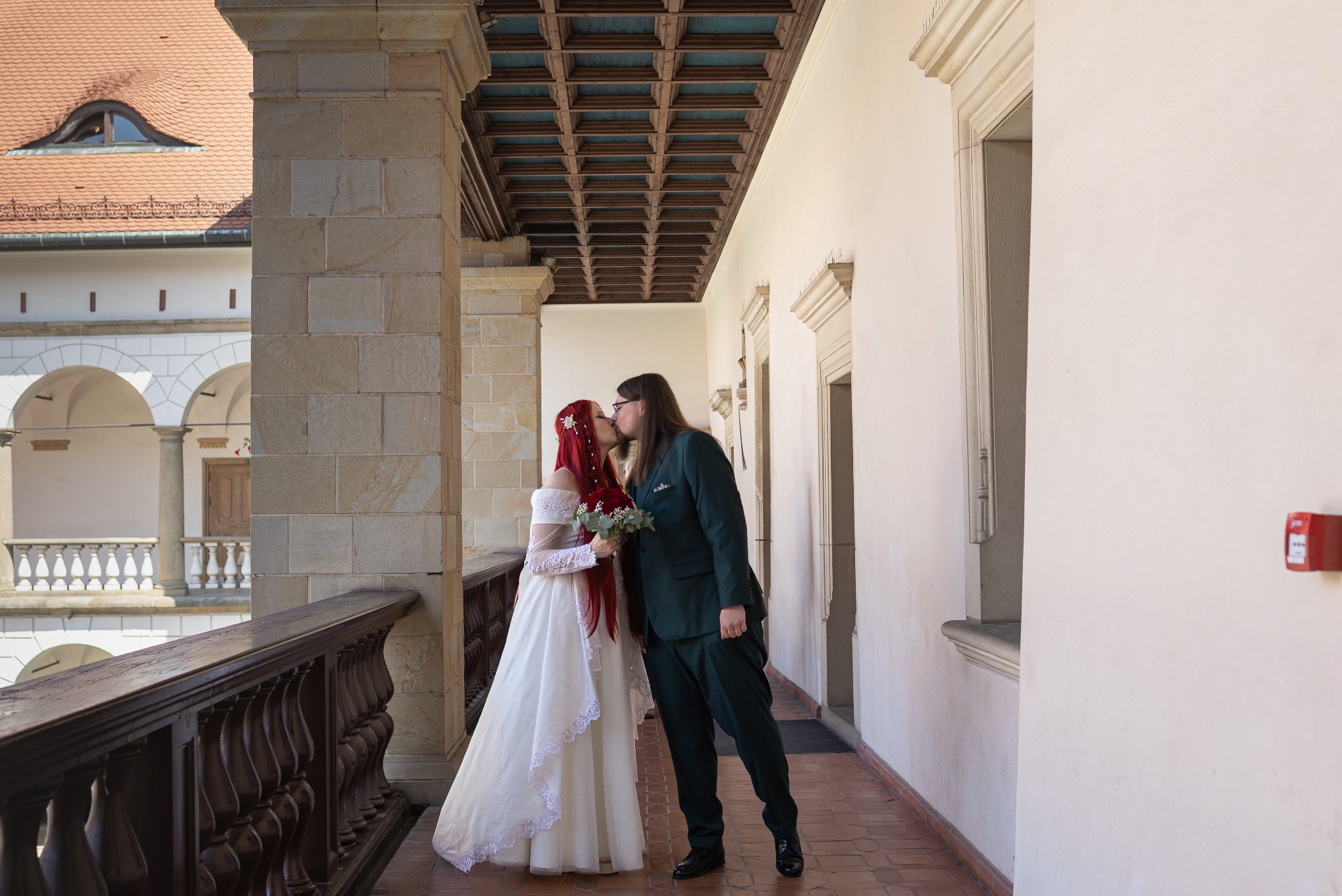 Pocałunek nowożeńców na tle zamkowego krużganku. Panna młoda w białej sukni z czerwonymi włosami, pan młody w ciemnozielonym garniturze. Kamienna architektura.