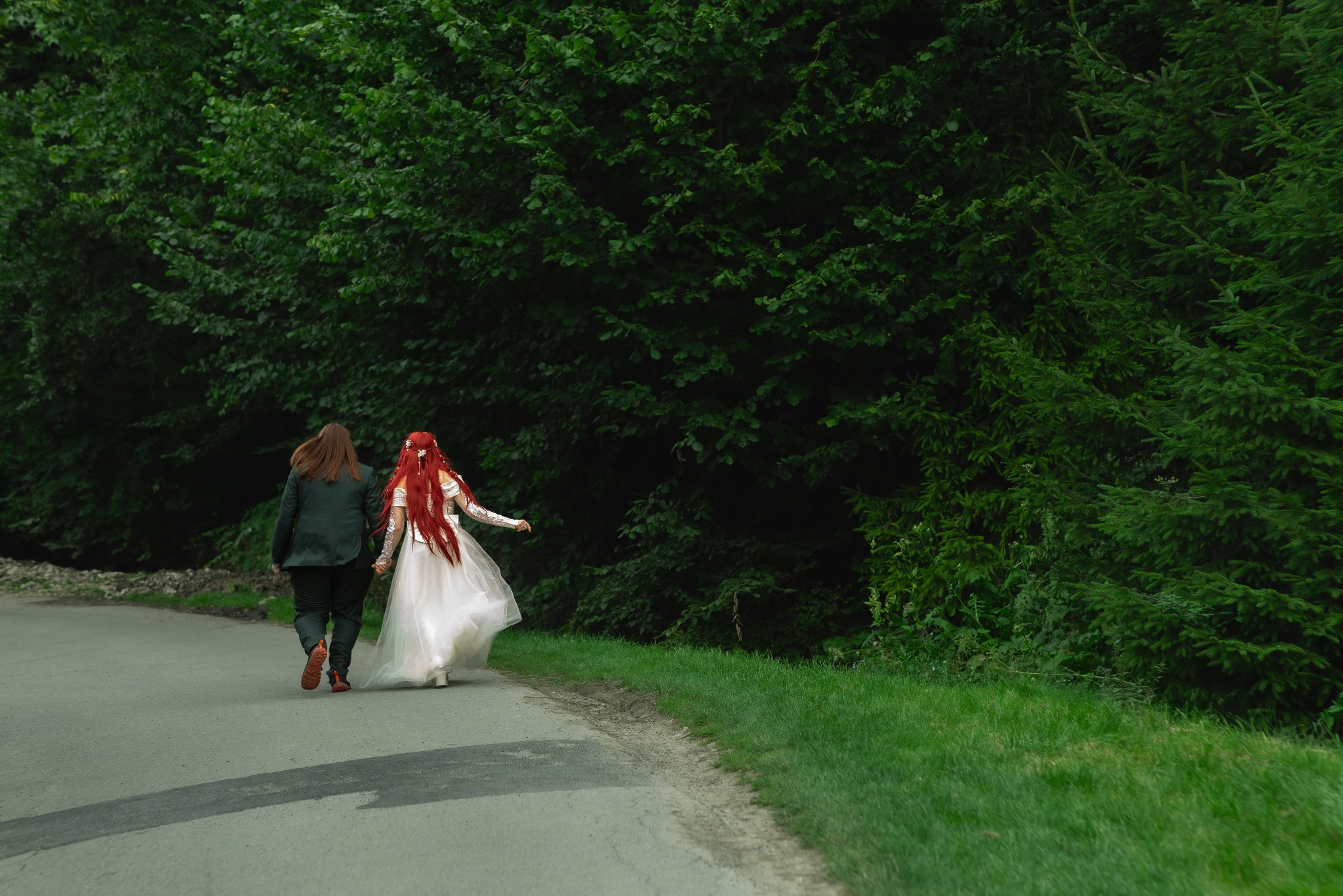 Para w stylizowanych strojach ślubnych, idąca leśną drogą. Panna młoda ma długie, czerwone włosy i białą suknię. Ujęcie z tyłu, w tle zielona roślinność.