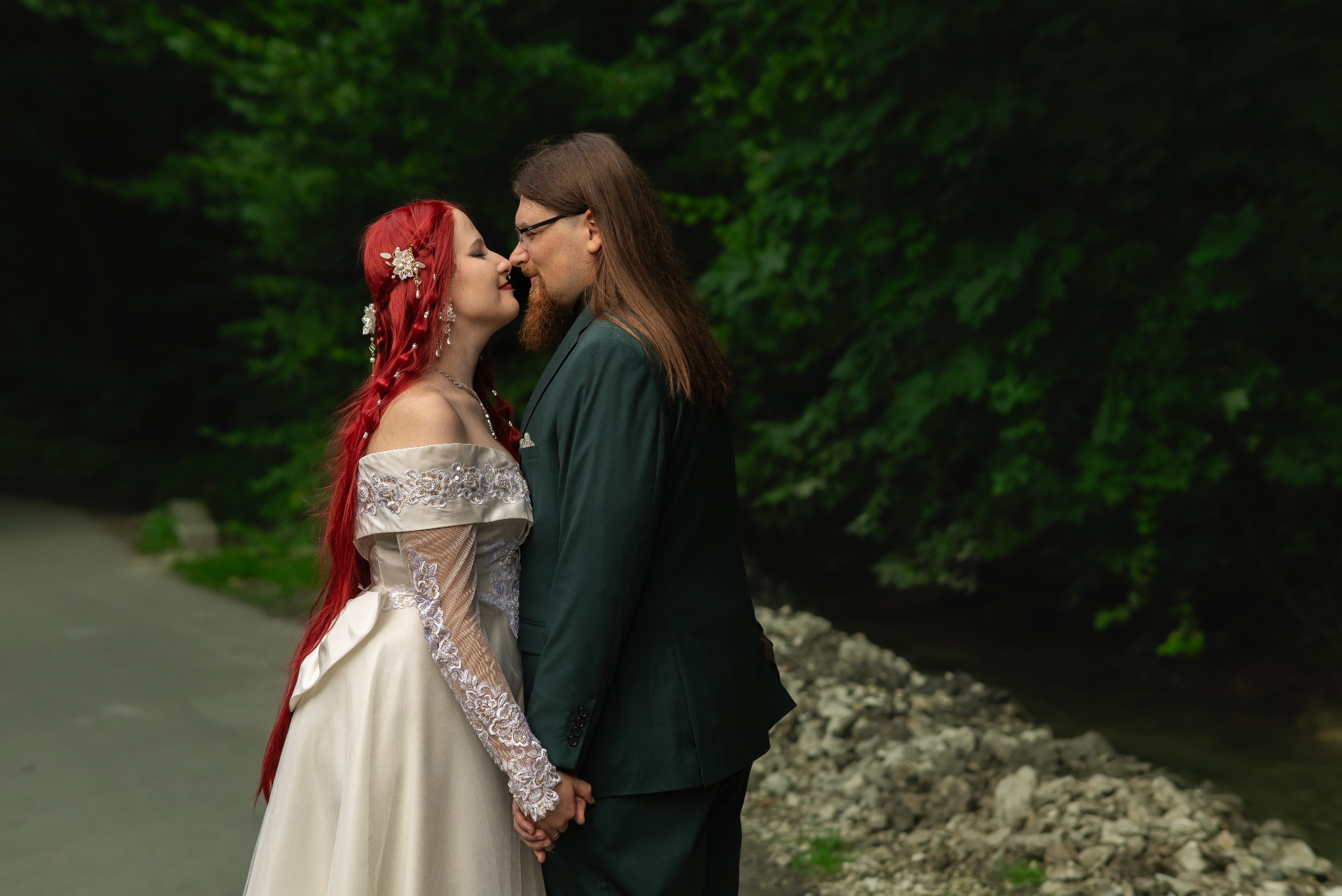 Para w strojach ślubnych, panna młoda z rudymi włosami i kwiatami, pan młody w zielonym garniturze, trzymają się za ręce na tle zieleni. Romantyczna sesja plenerowa.