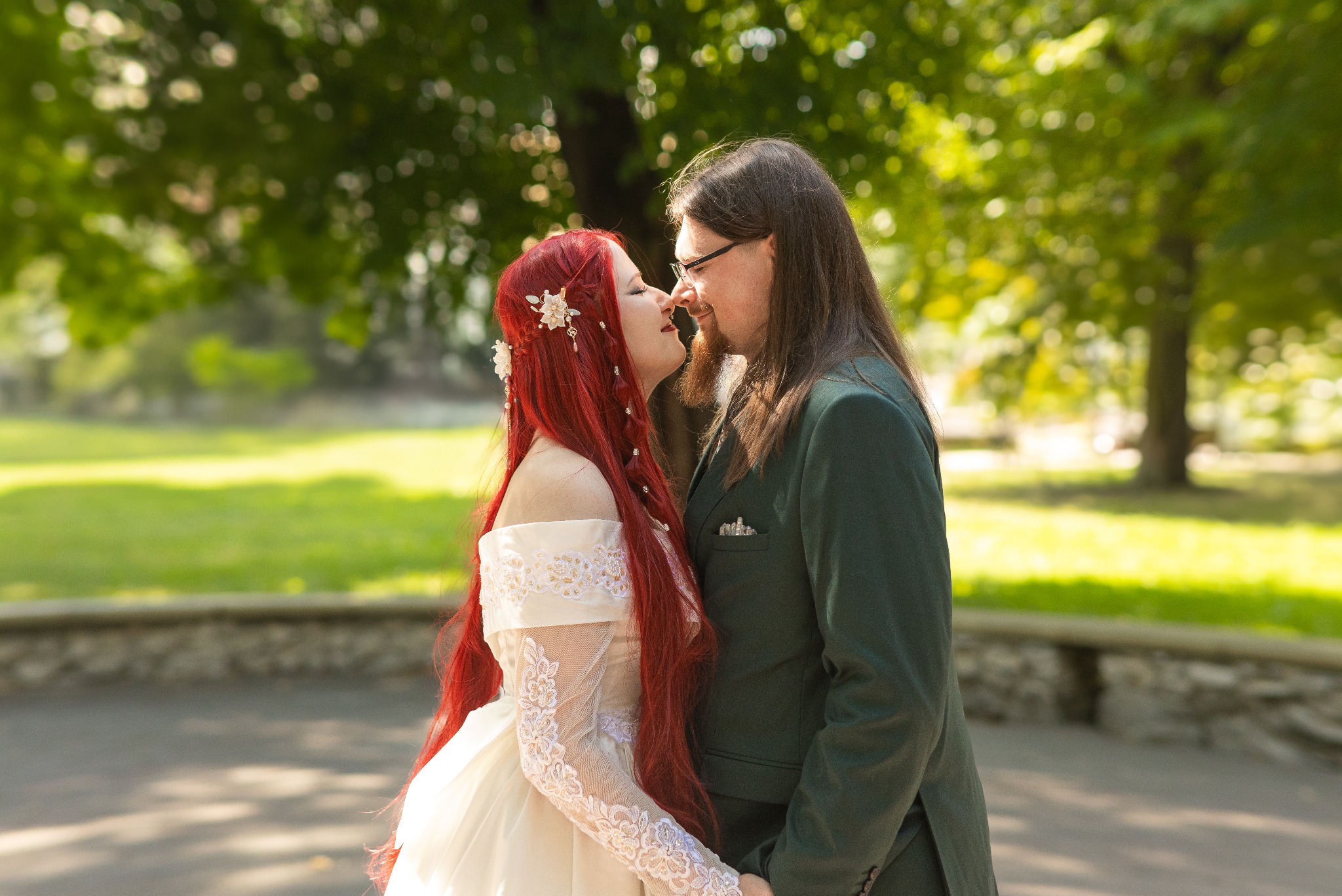 Para w strojach ślubnych, on w zielonym garniturze, ona w białej sukni z koronkowymi rękawami i czerwonymi włosami, delikatnie dotykają się nosami na tle parku.
