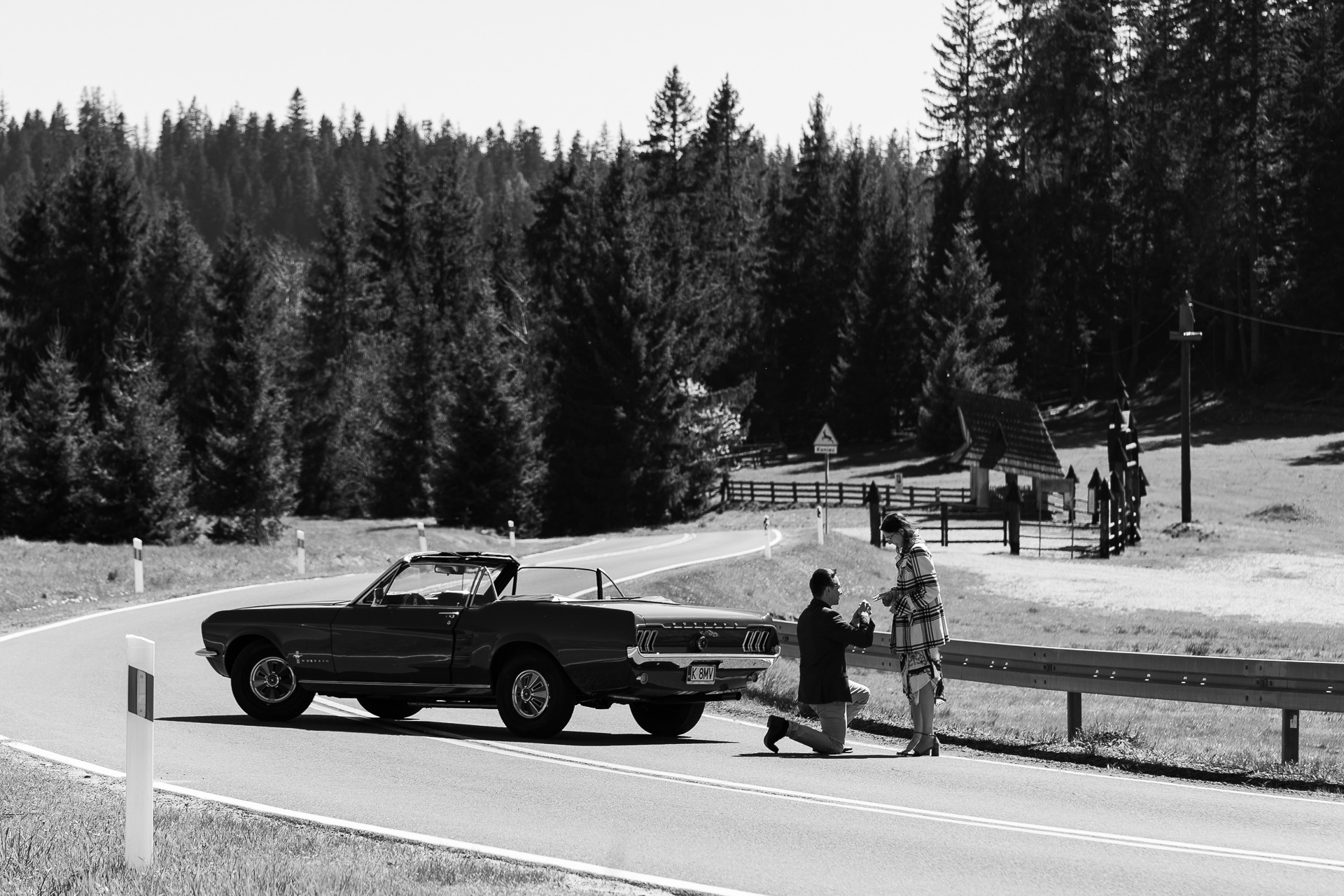 Czarno-biała fotografia zaręczyn na drodze z kabrioletem Mustang w tle, mężczyzna klęczy z pierścionkiem, w tle las i mały budynek.