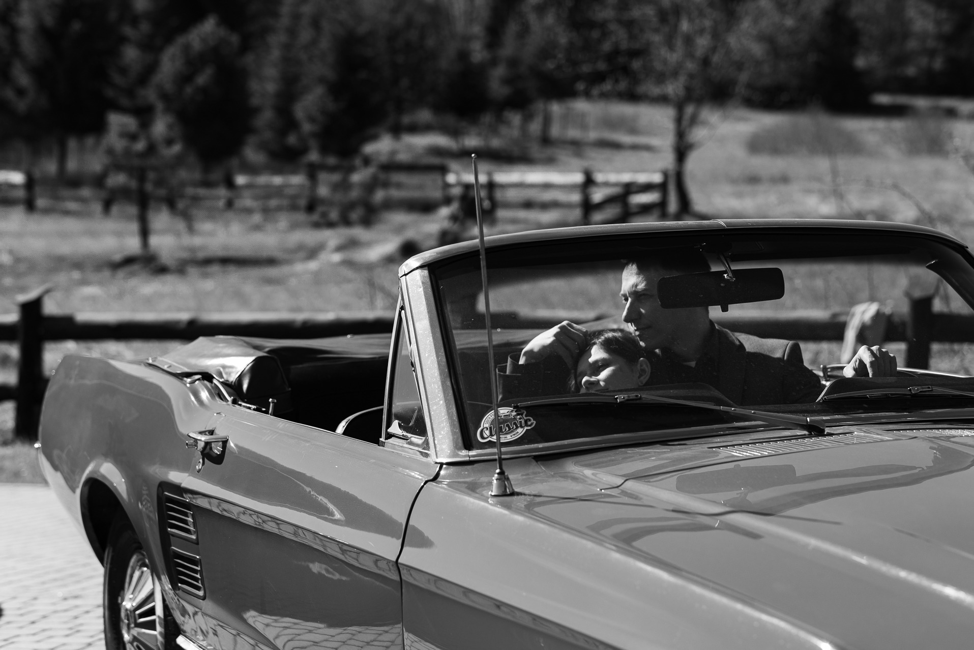 Czarno-białe zdjęcie pary w klasycznym kabriolecie Mustang w Krakowie. Ujęcie z zewnątrz, widoczny profil mężczyzny i fragment twarzy kobiety, tło lekko rozmyte.