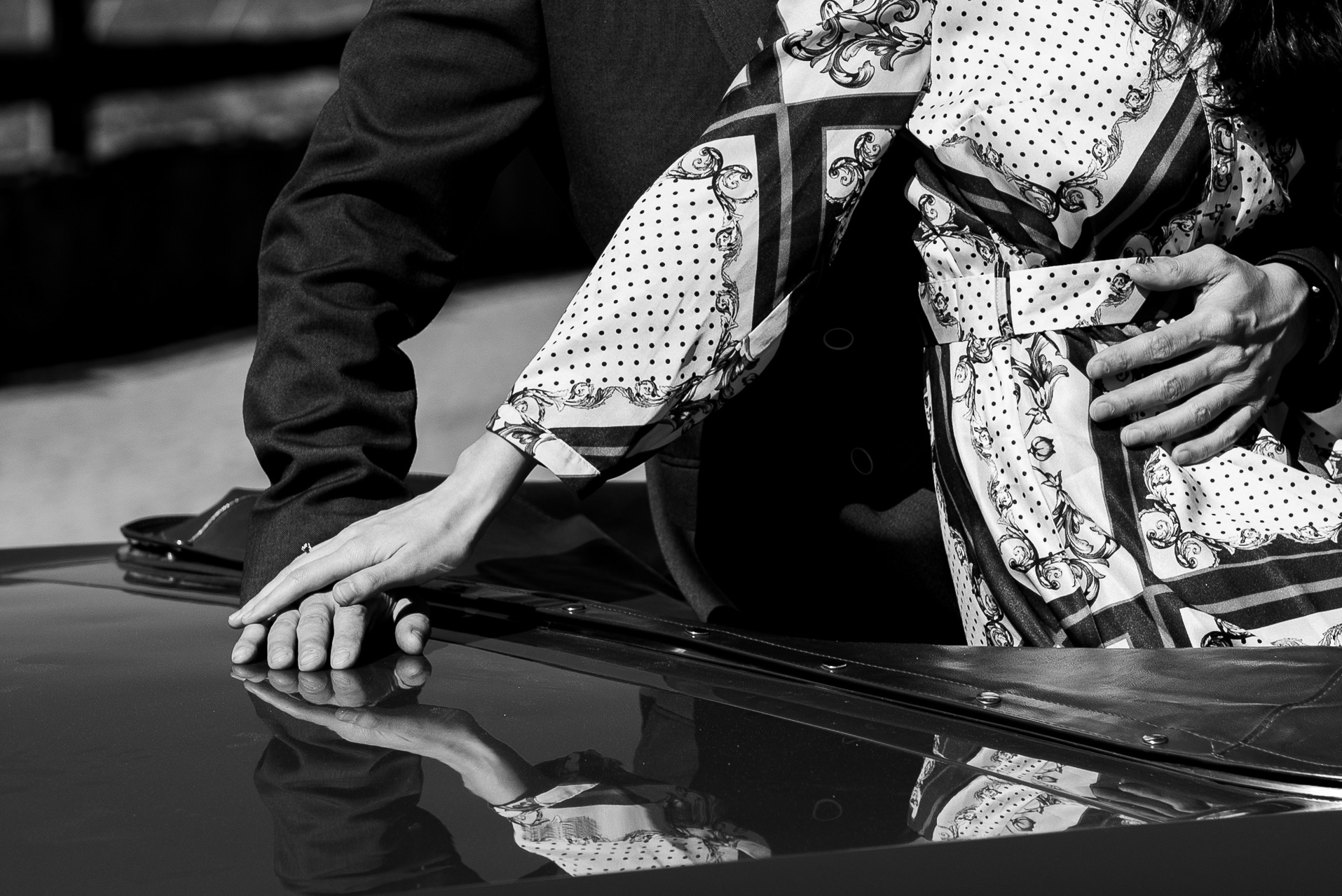 Czarno-białe zbliżenie dłoni pary na błyszczącej karoserii samochodu, odbicie sylwetek w lakierze. Ona w wzorzystej sukience, on w ciemnym garniturze.