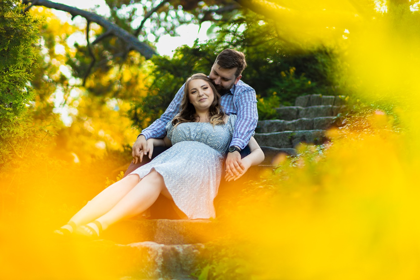 Zakochana para siedzi na kamiennych schodach w parku, mężczyzna obejmuje kobietę, ciepłe światło słoneczne, rozmyte żółte kwiaty w pierwszym planie.