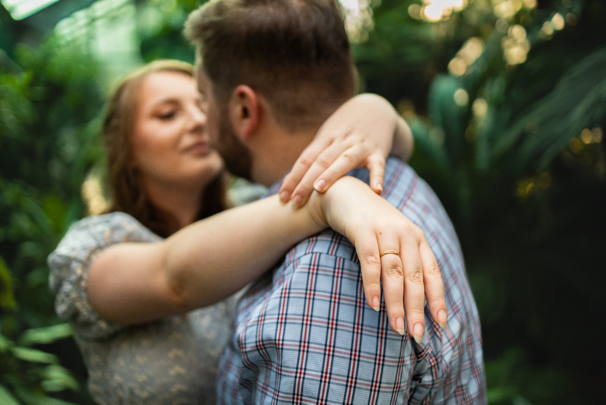Para w objęciach w szklarni, kobieta z pierścionkiem na palcu obejmuje mężczyznę, rozmyte tło zieleni. Romantyczna chwila, ciepłe światło.