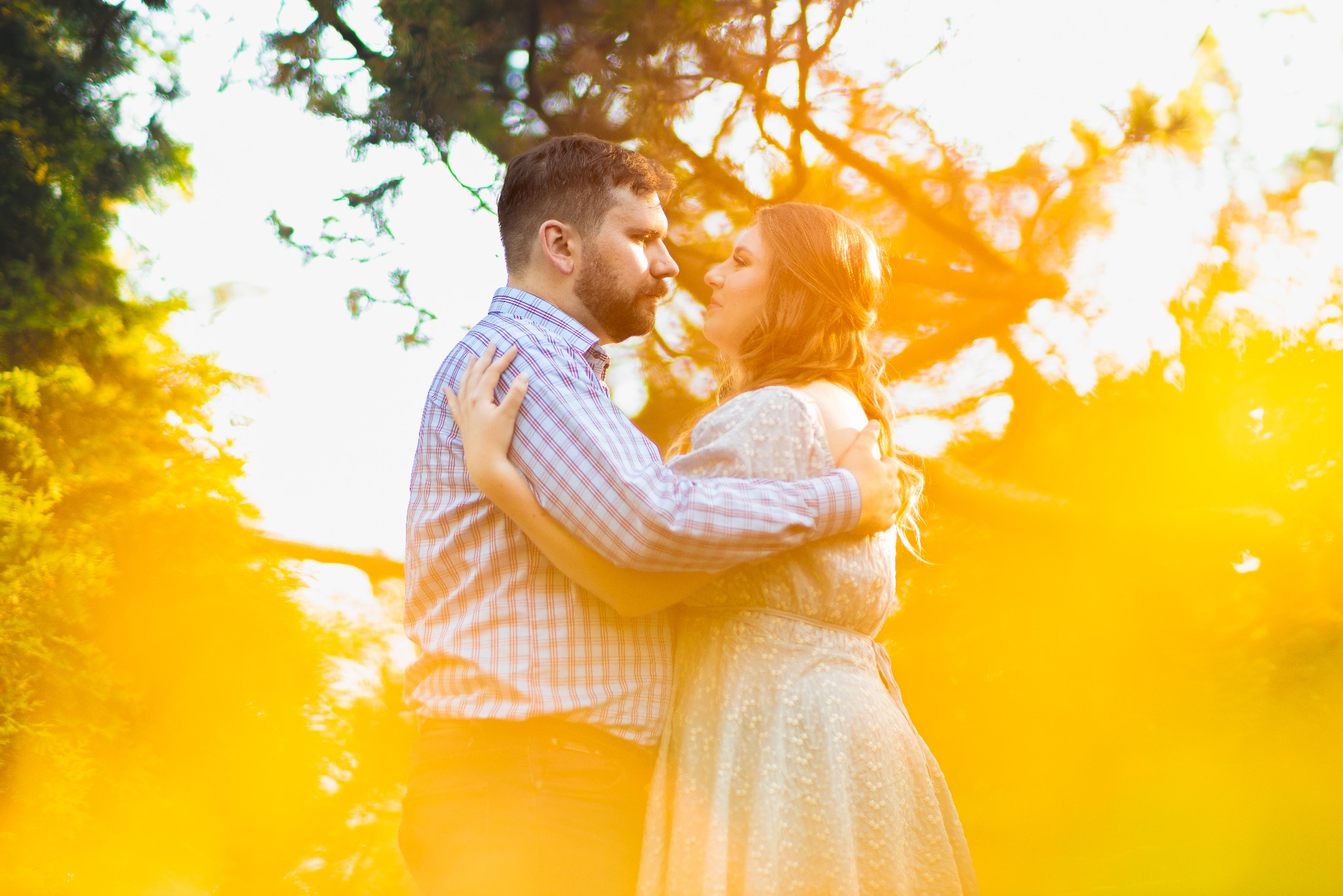 Zakochana para w plenerze, obejmująca się w kontraście z promieniami słońca. Kobieta w jasnej sukience, mężczyzna w koszuli w kratę. Romantyczna atmosfera.