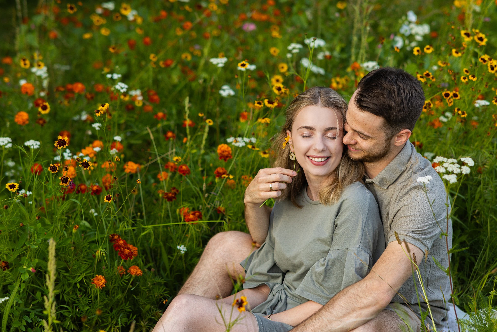 Zakochana para siedzi w kwitnącej łące. Kobieta z zamkniętymi oczami trzyma kwiatek przy uchu, uśmiechając się. Mężczyzna obejmuje ją czule, wtulony w jej włosy.