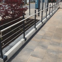 Magia Metalu Krzysztof Muzolf - Nowoczesna balustrada z metalowymi słupkami i poziomymi linkami, obok rabaty z czerwonym klonem palmowym, na tle jasnej kostki brukowej i elementów infrastruktury miejskiej.