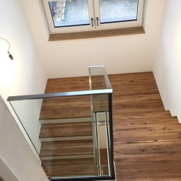 Magia Metalu Krzysztof Muzolf - Nowoczesne schody drewniane z balustradą ze szkła, widok z góry. Naturalne oświetlenie z okna na poddaszu. Minimalistyczny design, ciepłe odcienie drewna.