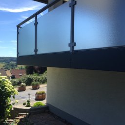 Magia Metalu Krzysztof Muzolf - Szklana balustrada balkonu z ciemnoszarą ramą i widokiem na okolicę w słoneczny dzień. Charakterystyczne mocowania szkła.