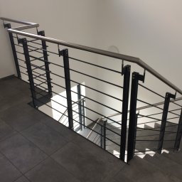 Magia Metalu Krzysztof Muzolf - Metalowe schody z balustradą o konstrukcji z czarnych słupków i poziomych prętów, z górną poręczą w kolorze srebrnym. Widok z góry, z gładką szarą podłogą.