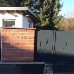 Nowoczesna brama wjazdowa z metalu w kolorze grafitowym, zdobiona motywem roślinnym w postaci wyciętych kłosów zboża, osadzona między ceglanymi i kamiennymi słupkami.