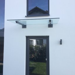 Magia Metalu Krzysztof Muzolf - Szklany daszek nad wejściem do domu z grafitową stolarką okienną i drzwiową. Nowoczesny design z widocznymi mocowaniami. Minimalizm i funkcjonalność.