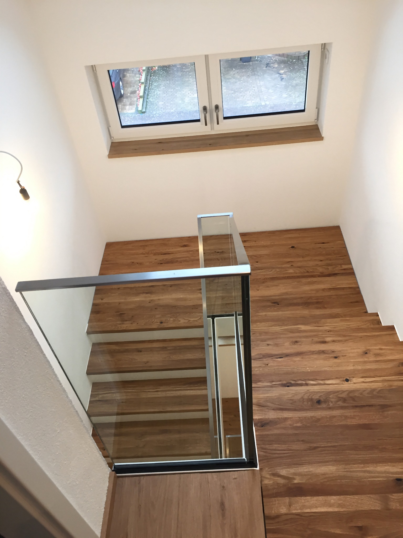 Nowoczesne schody drewniane z balustradą ze szkła, widok z góry. Naturalne oświetlenie z okna na poddaszu. Minimalistyczny design, ciepłe odcienie drewna.