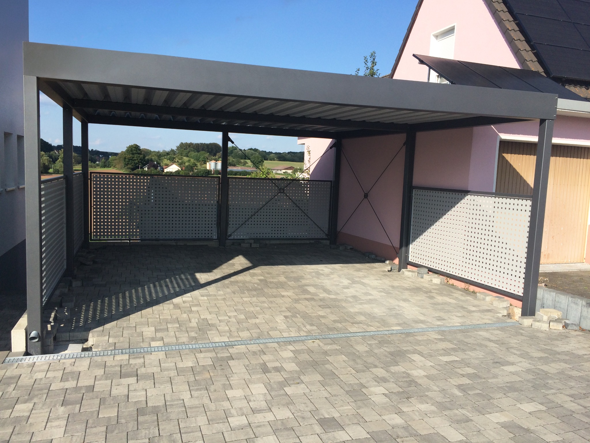 Nowoczesna, szara wiata garażowa z perforowanymi panelami bocznymi, na tle różowego domu z panelami słonecznymi i brukowanym podjazdem.