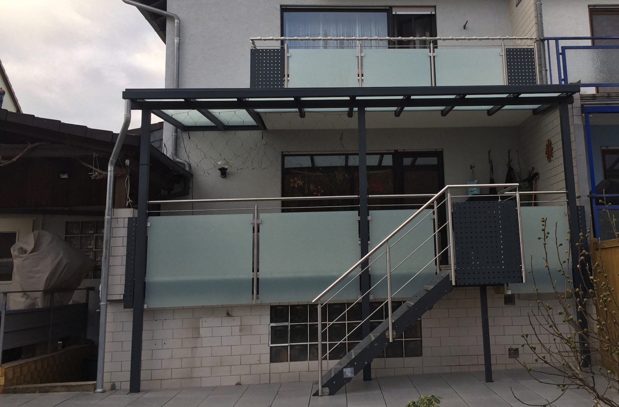 Zadaszony balkon z metalowymi schodami i szklanymi balustradami, widok z dołu. Metalowa konstrukcja w kolorze grafitowym i srebrne poręcze.