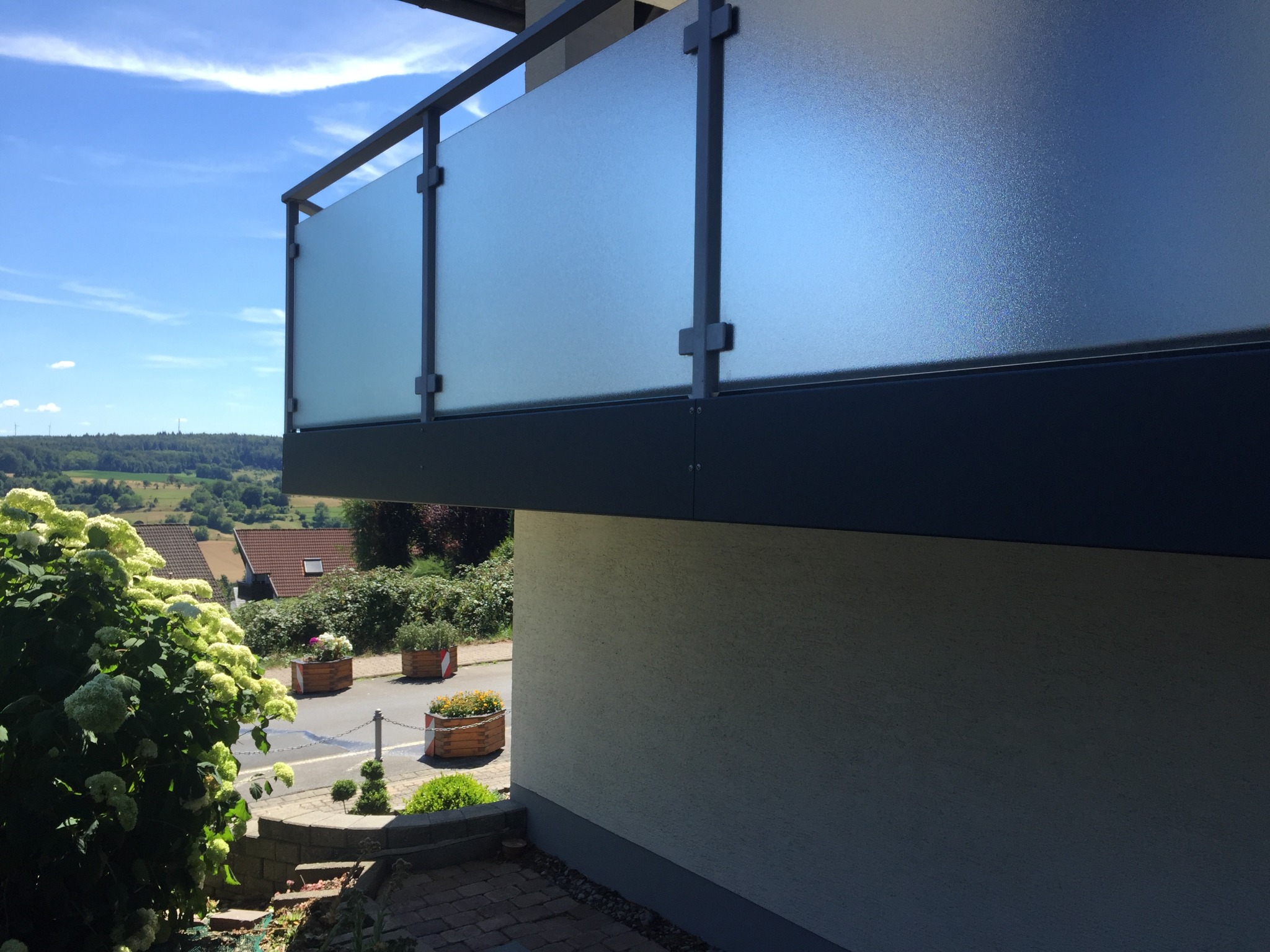 Szklana balustrada balkonu z ciemnoszarą ramą i widokiem na okolicę w słoneczny dzień. Charakterystyczne mocowania szkła.