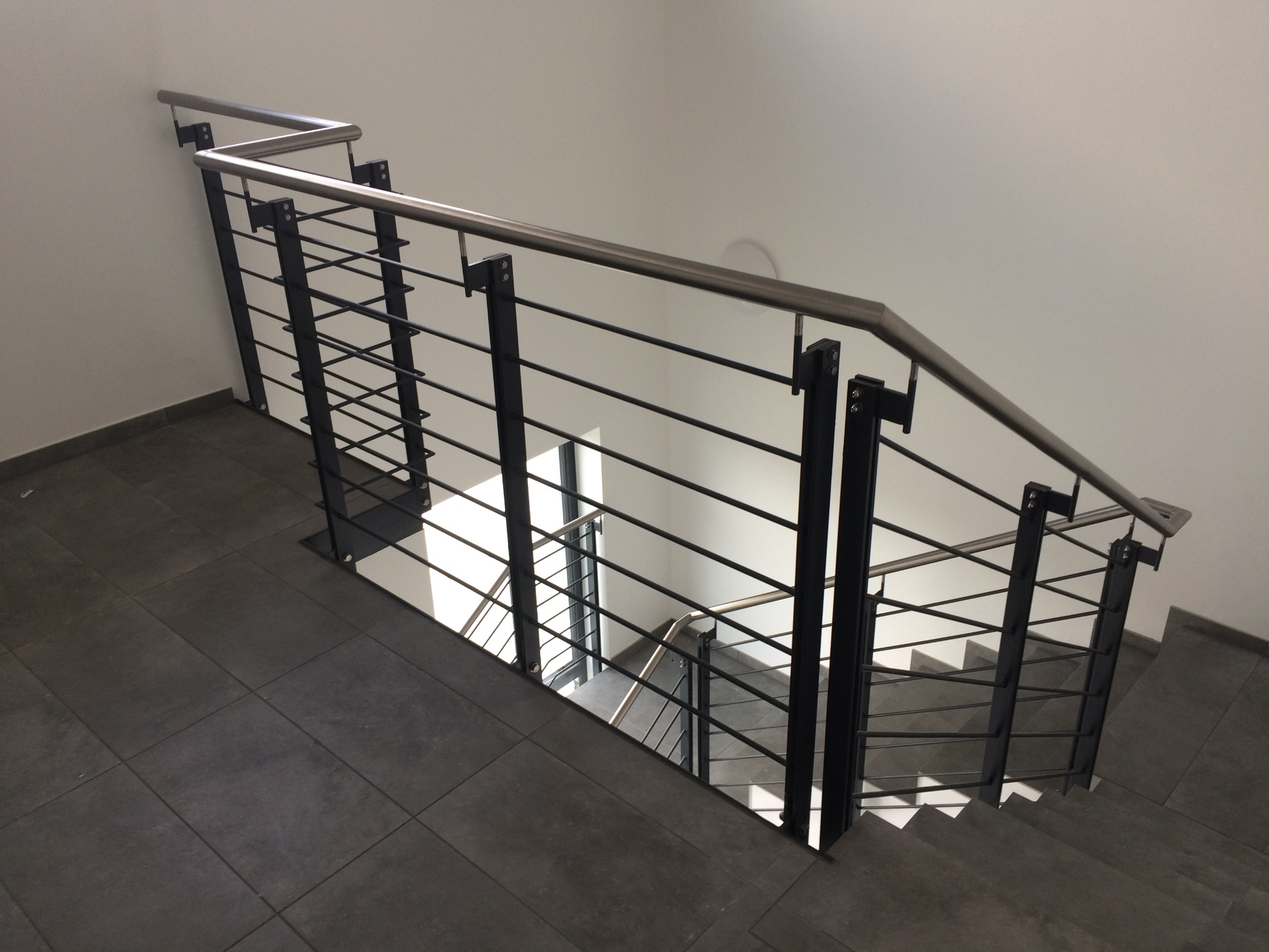 Metalowe schody z balustradą o konstrukcji z czarnych słupków i poziomych prętów, z górną poręczą w kolorze srebrnym. Widok z góry, z gładką szarą podłogą.