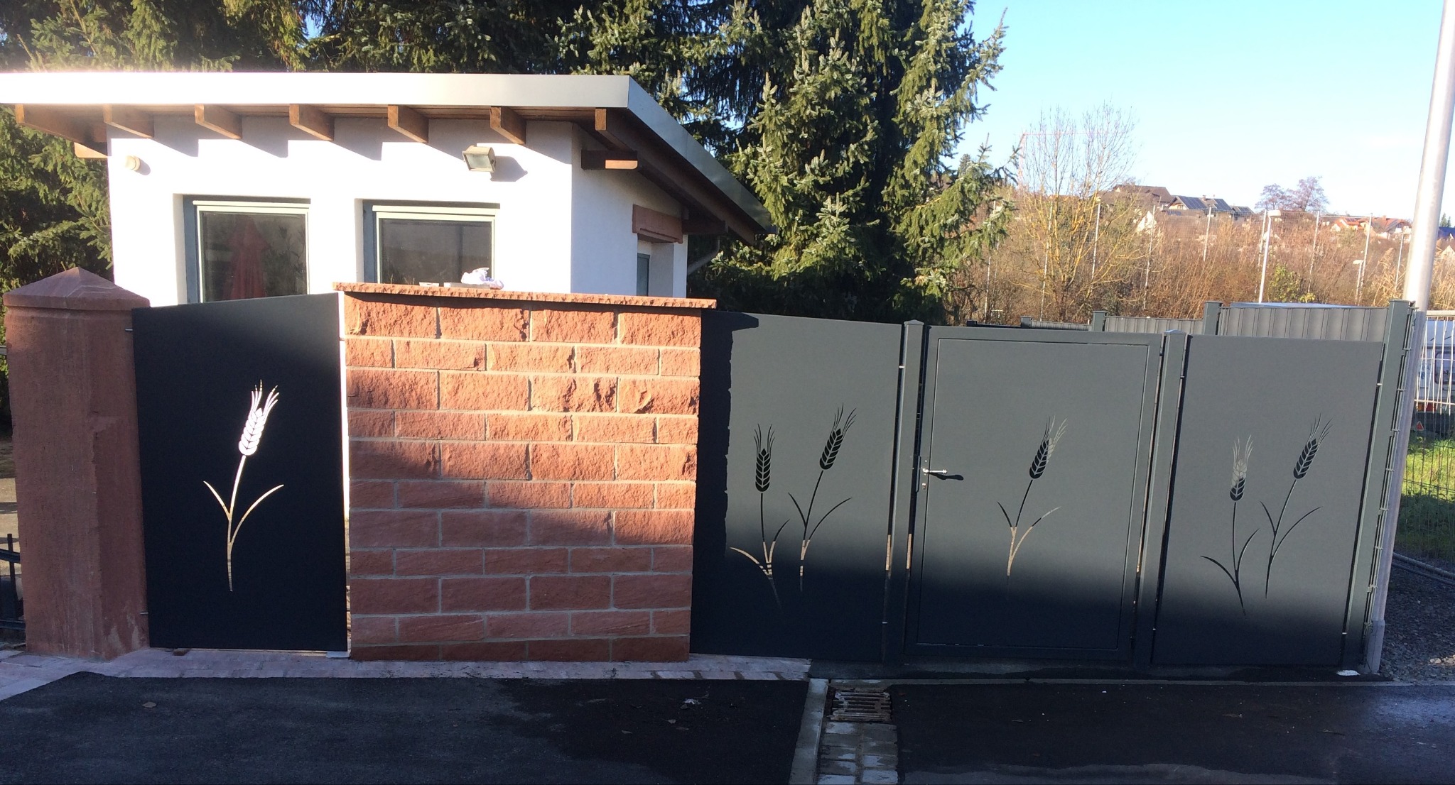 Nowoczesna brama wjazdowa z metalu w kolorze grafitowym, zdobiona motywem roślinnym w postaci wyciętych kłosów zboża, osadzona między ceglanymi i kamiennymi słupkami.