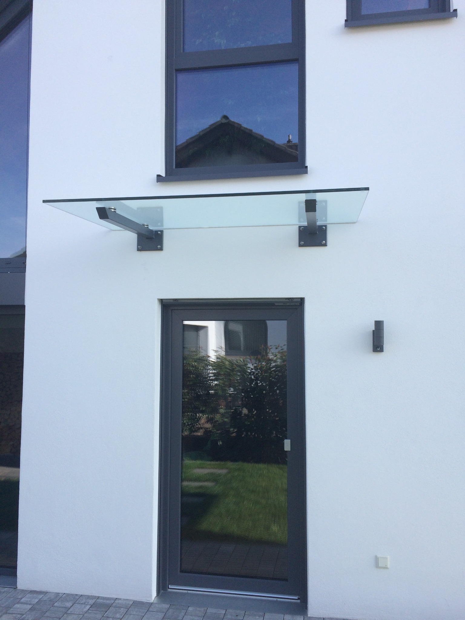 Szklany daszek nad wejściem do domu z grafitową stolarką okienną i drzwiową. Nowoczesny design z widocznymi mocowaniami. Minimalizm i funkcjonalność.