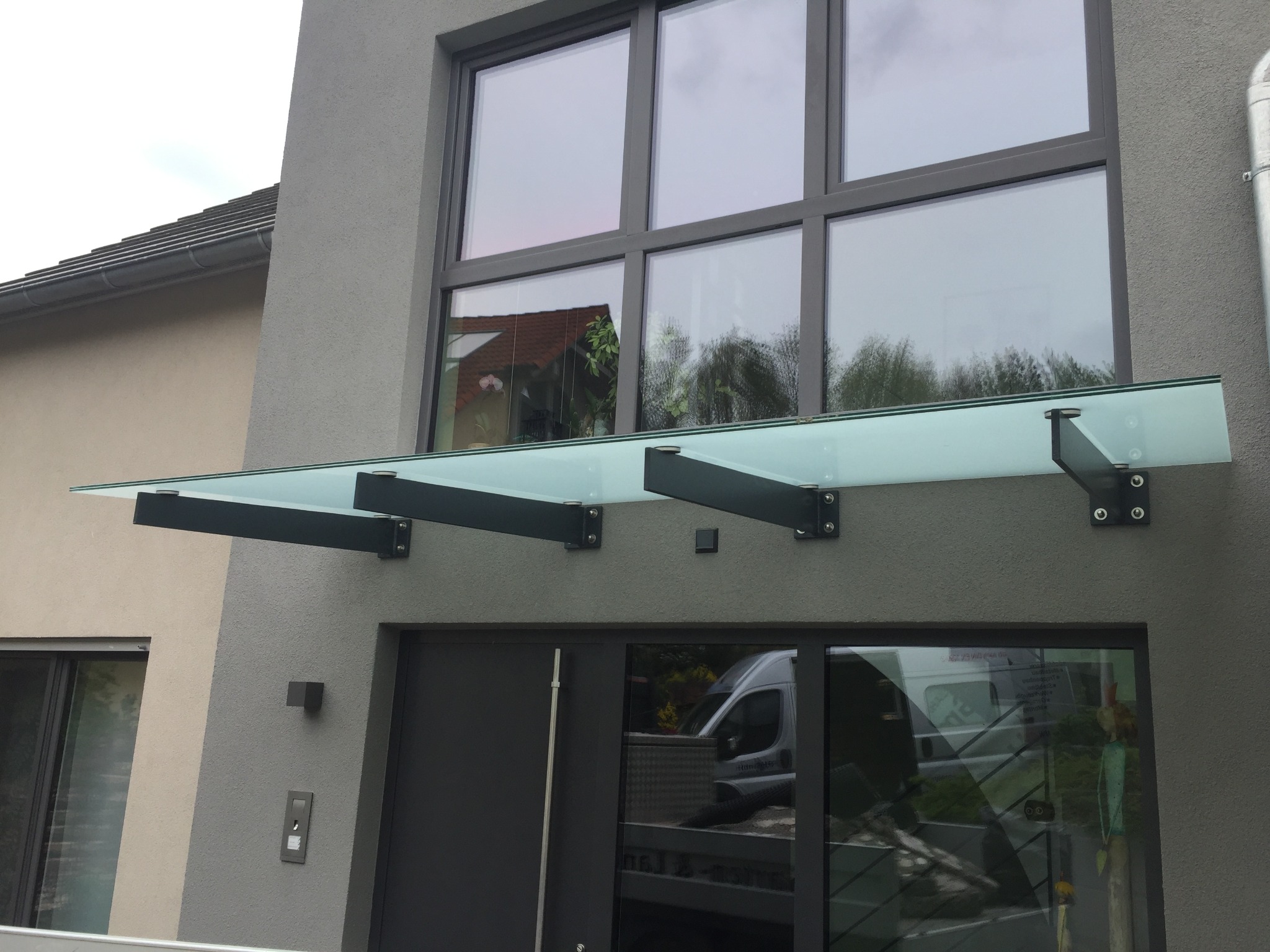 Szklany daszek nad nowoczesnym wejściem do domu z szarą elewacją. Konstrukcja wsparta na czarnych, metalowych wspornikach. Widoczne duże okno i drzwi.