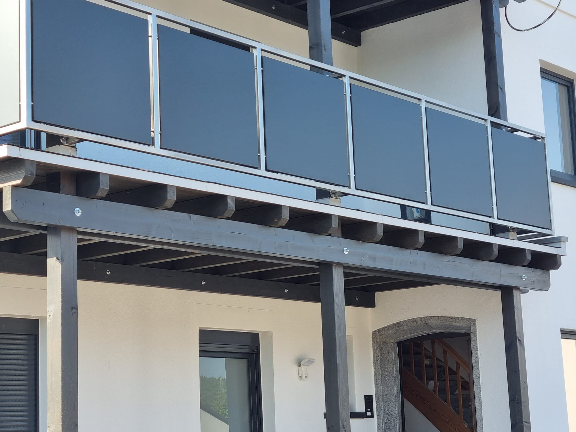 Nowoczesna balustrada szklana z metalowymi elementami na balkonie z drewnianą konstrukcją, widok z dołu, jasne tło budynku.