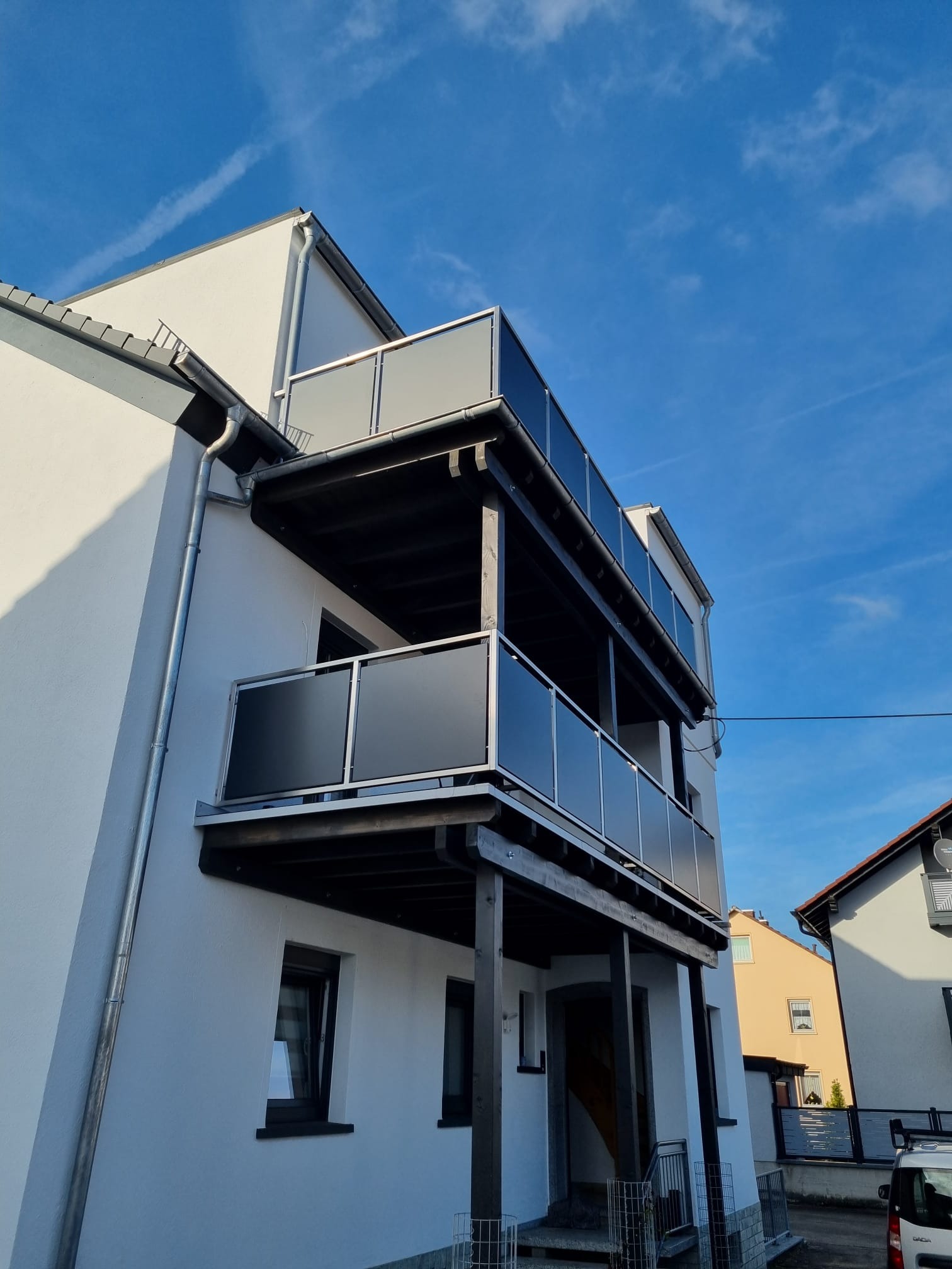 Dwupoziomowy balkon z grafitowymi szklanymi balustradami i ciemnym drewnem, widziany z dołu pod kątem, na tle błękitnego nieba z delikatnymi smugami chmur.