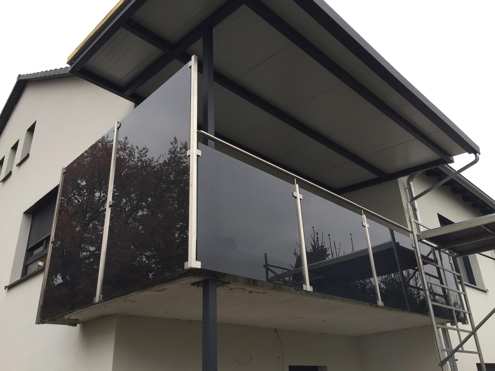 Nowoczesna balustrada szklana balkonu z metalowymi słupkami i poręczą, odbijająca otoczenie, na tle elewacji domu z szarym dachem. Widoczne rusztowanie.