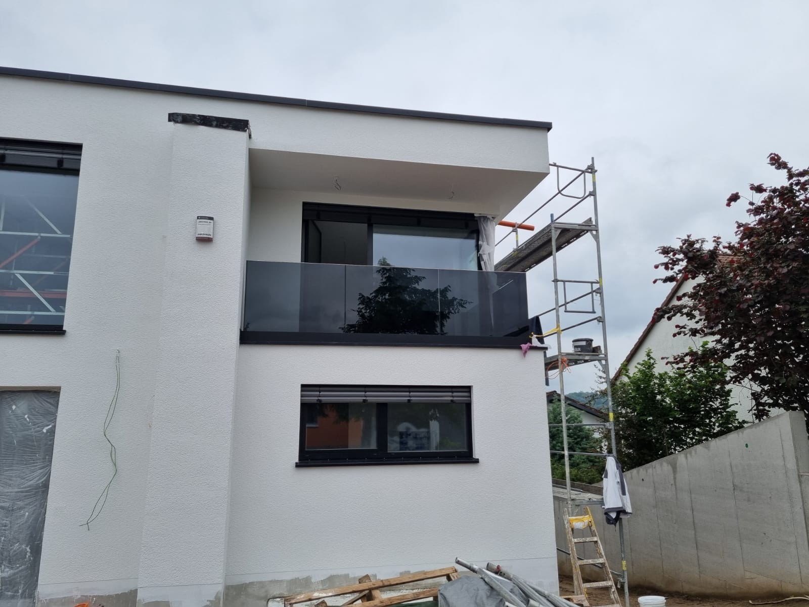 Nowoczesny budynek z balkonem zabezpieczonym szklaną balustradą w czarnej ramie. Widoczne rusztowanie i elementy wykończeniowe.