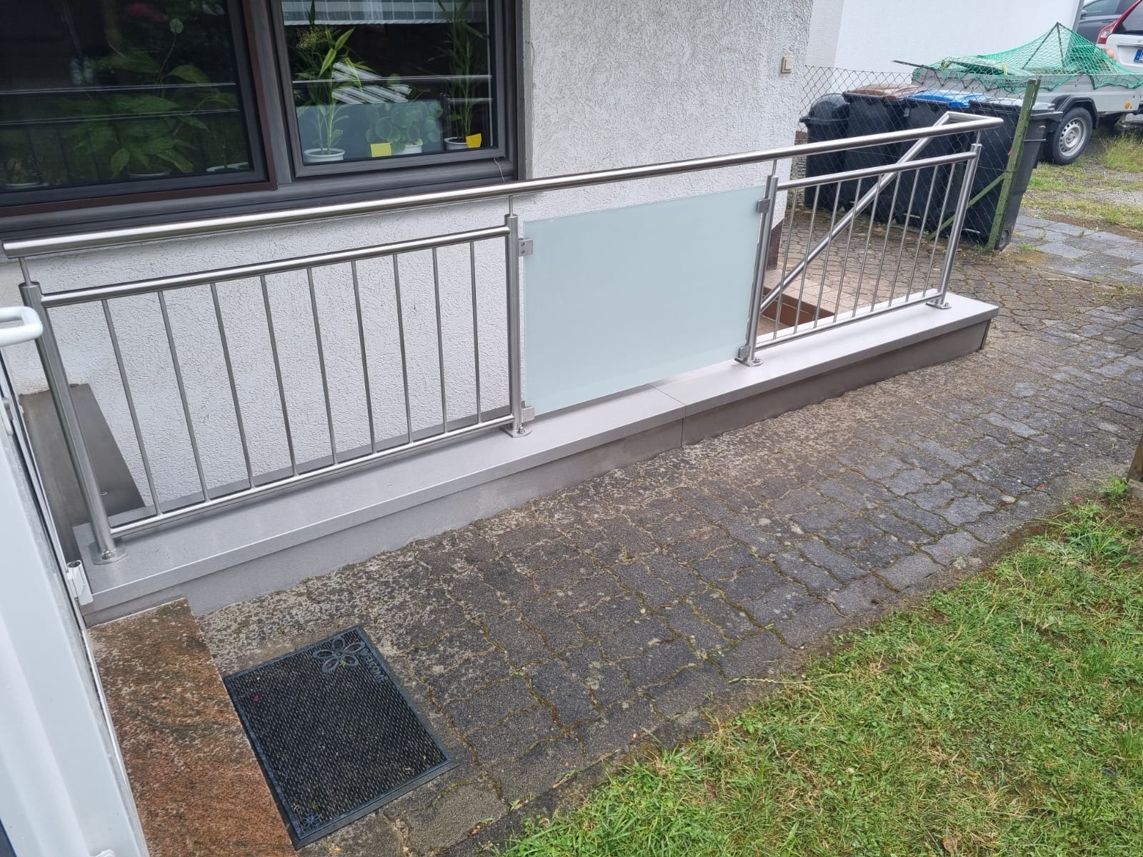 Nowoczesna balustrada ze stali nierdzewnej i szkła matowego, zamontowana przy wejściu do domu. Widoczne detale wykończenia i otoczenie budynku.