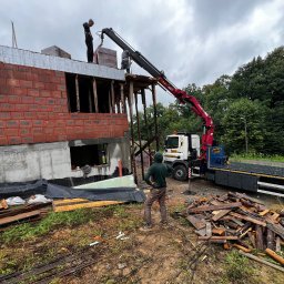 JAROSŁAW TABOŁA TAB-HOL HDS - Budowa domu z czerwonej cegły. Żuraw przenosi materiały na górę. Widoczny pracownik w zielonej bluzie i laweta. Tło: las i pochmurne niebo.