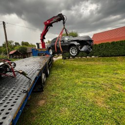 JAROSŁAW TABOŁA TAB-HOL HDS - Czarny, uszkodzony samochód podnoszony przez czerwony dźwig lawety na tle pochmurnego nieba i zielonego trawnika. Widoczna część platformy lawety.