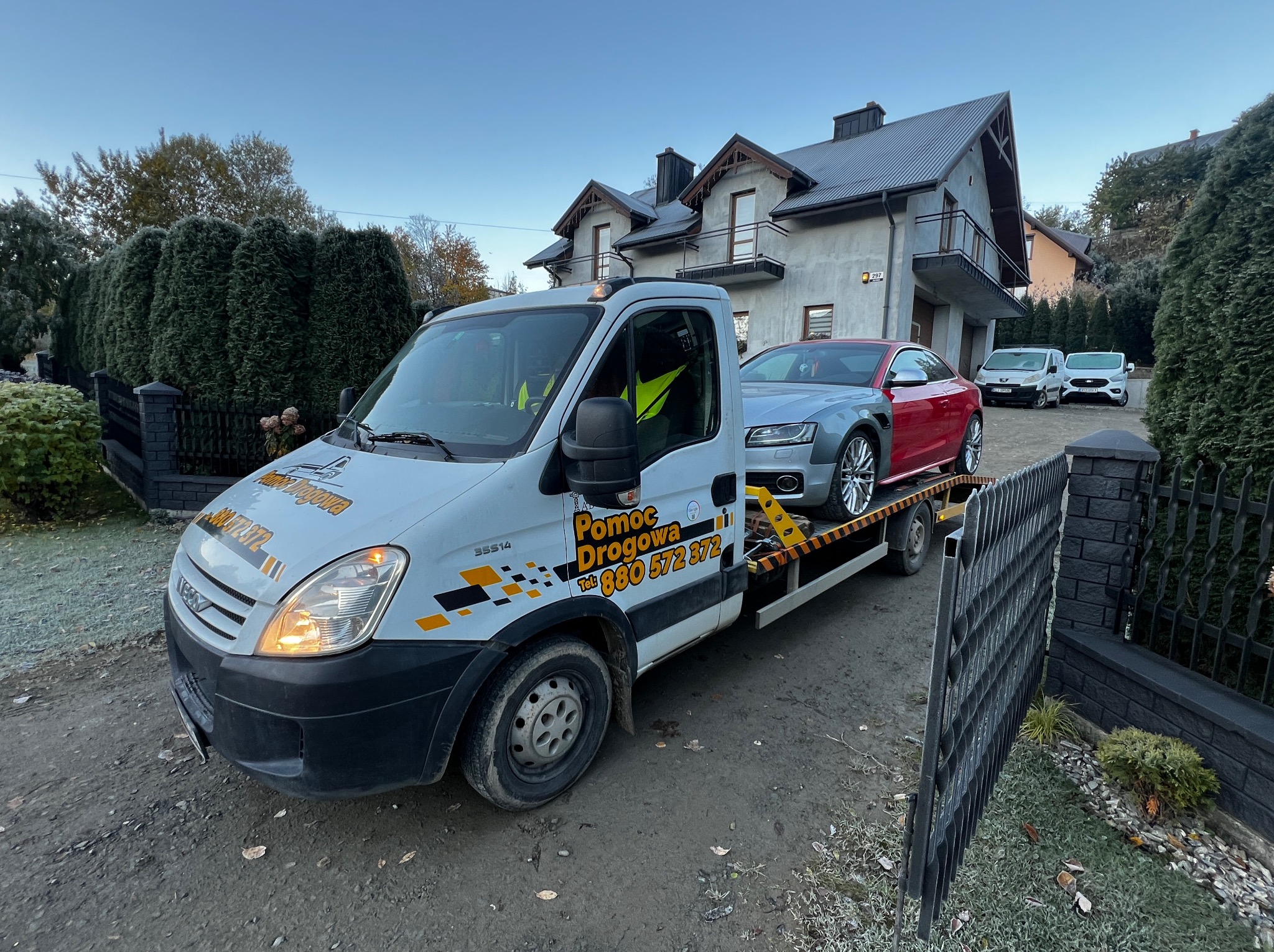 Laweta z Audi A5 na platformie, zaparkowana przed domem. Na białej kabinie widoczne logo 'Pomoc Drogowa' i numer telefonu. W tle inne pojazdy i budynek mieszkalny.