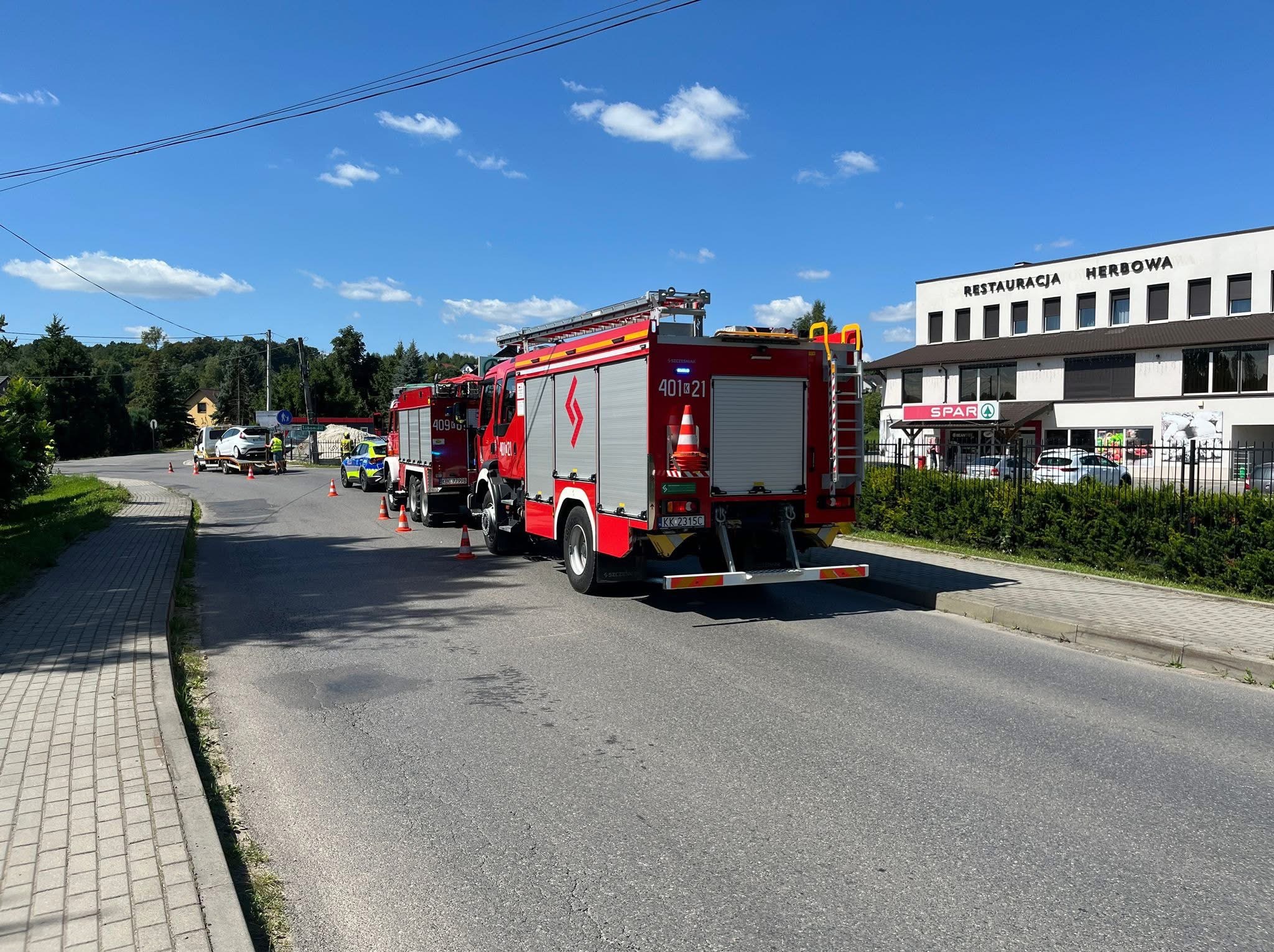 Dwa wozy strażackie na drodze, w tle auto na lawecie i radiowóz. Akcja ratownicza w słoneczny dzień, widoczny budynek z napisem 'Restauracja Herbowa'.