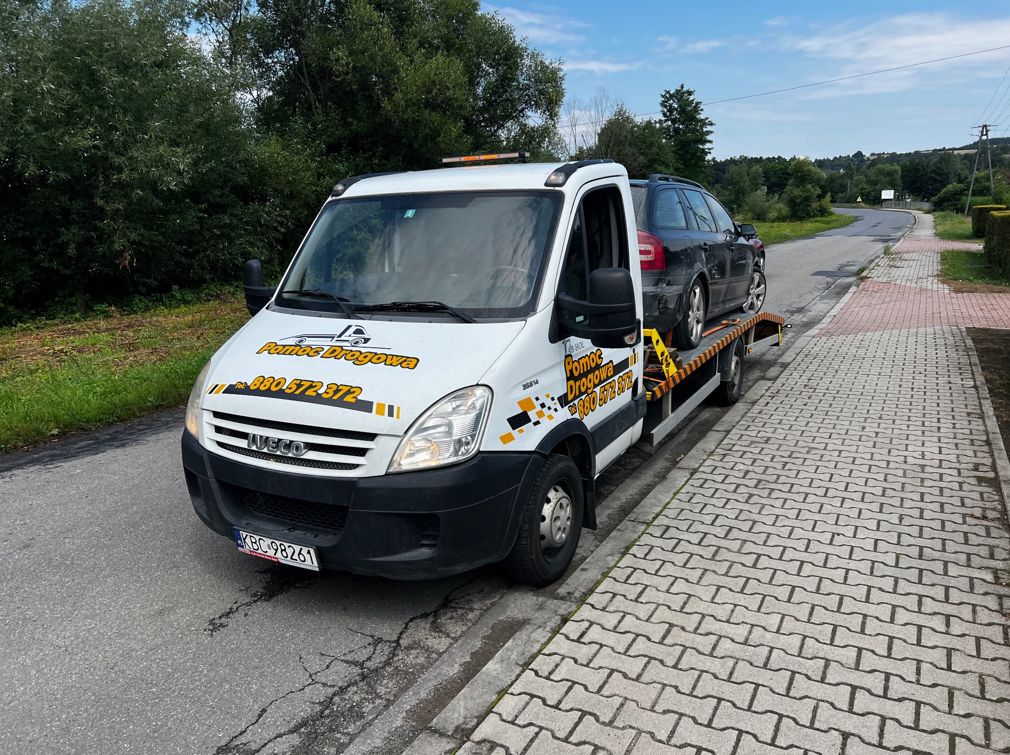 Laweta Iveco z czarnym samochodem osobowym na platformie, zaparkowana na poboczu drogi. Widoczne logo 'Pomoc Drogowa' i numer telefonu. Słoneczny dzień.